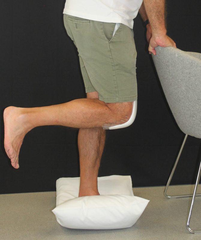 man standing on one leg on a pillow and holding onto a chair in front